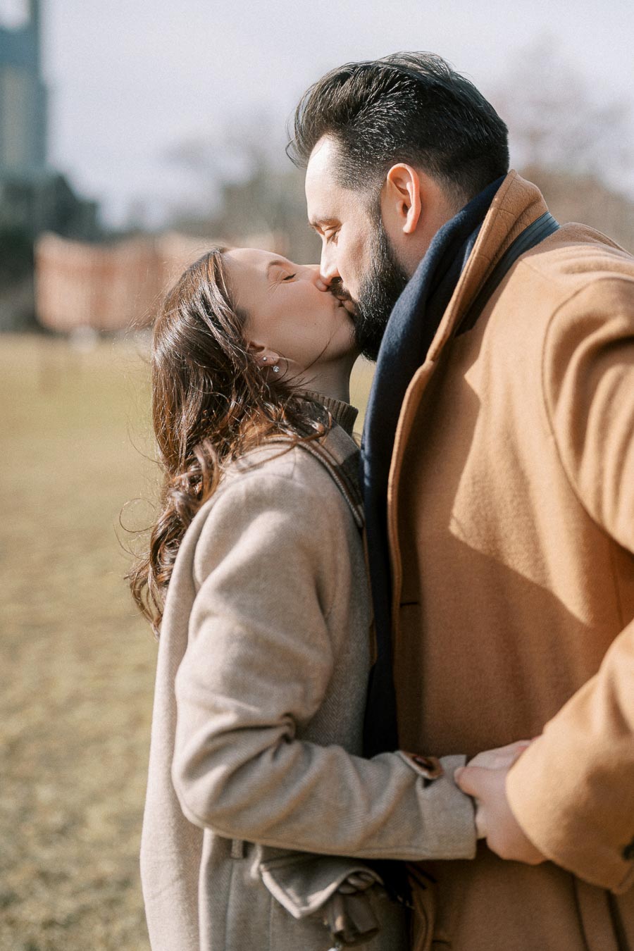 A couple in warm winter coats kissing outdoors on a sunny day, showcasing romance and affection.