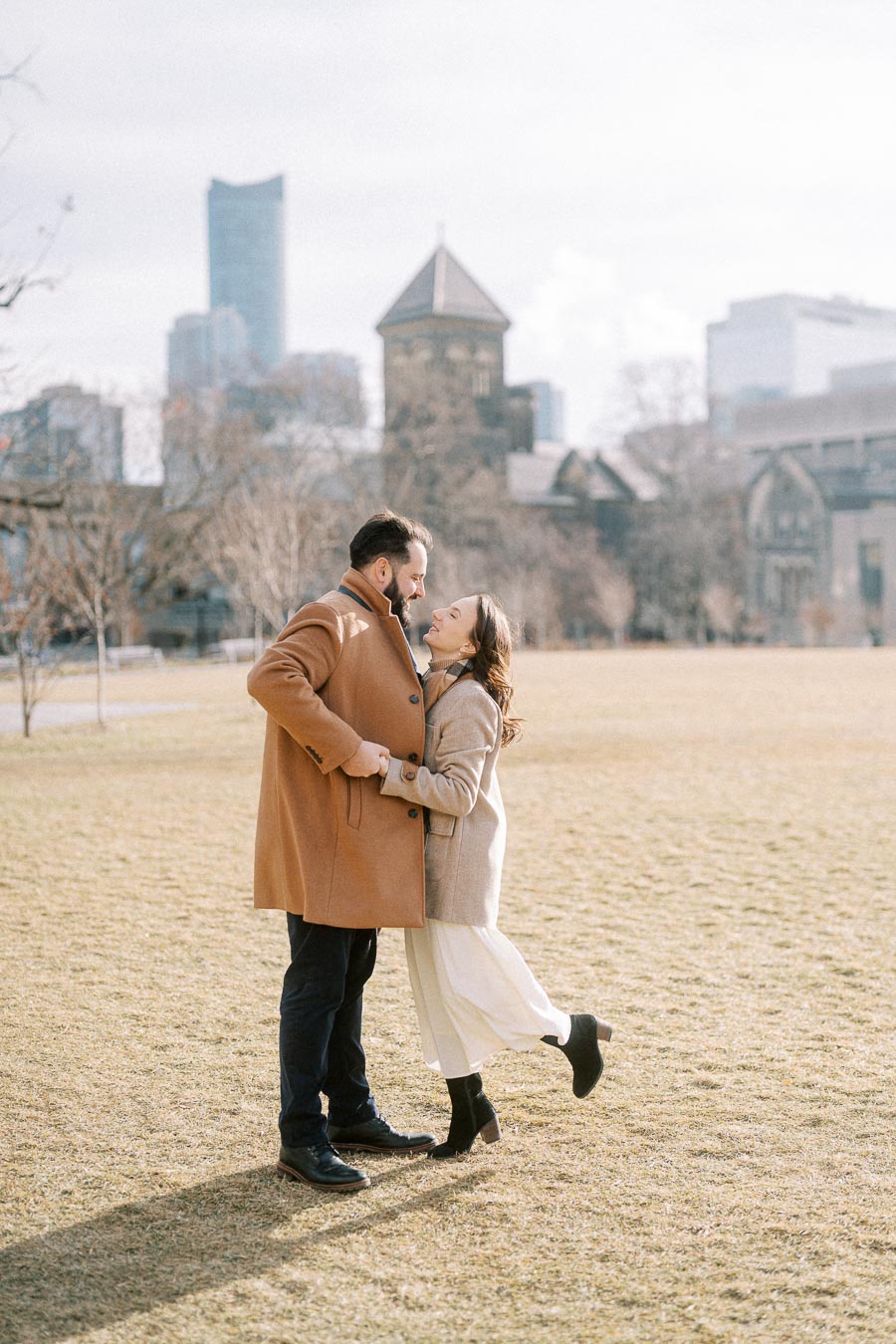 A couple in stylish winter coats embraces in a sunny park with city skyline in the background.