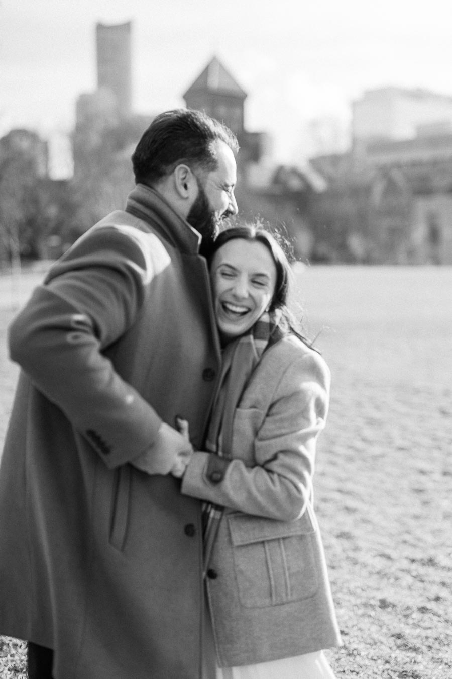 A black and white image of a couple joyfully embracing in a park, dressed in warm coats, with a cityscape in the blurred background.