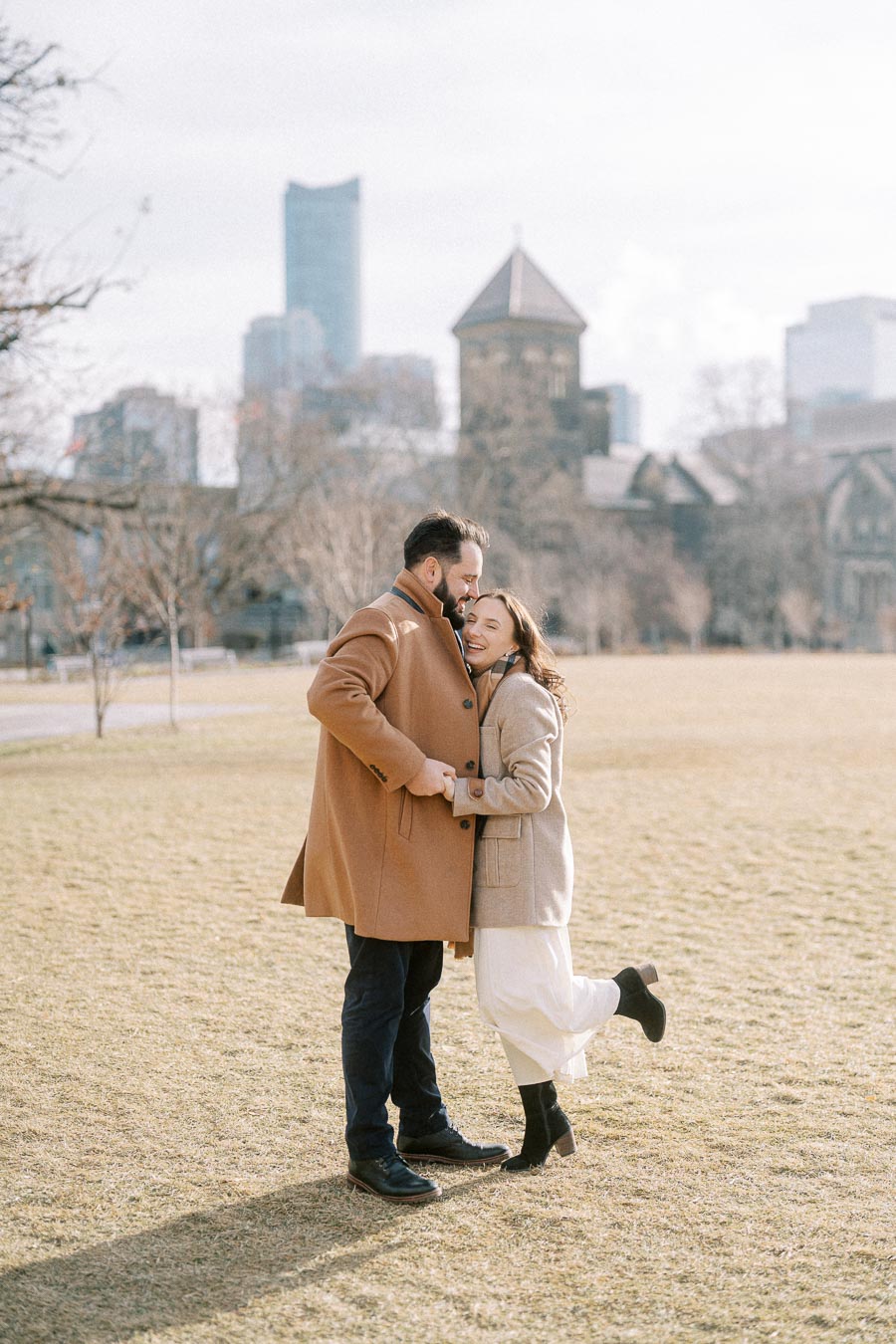 A couple in winter coats embracing in a sunny park, with city skyline and historic building in the background.