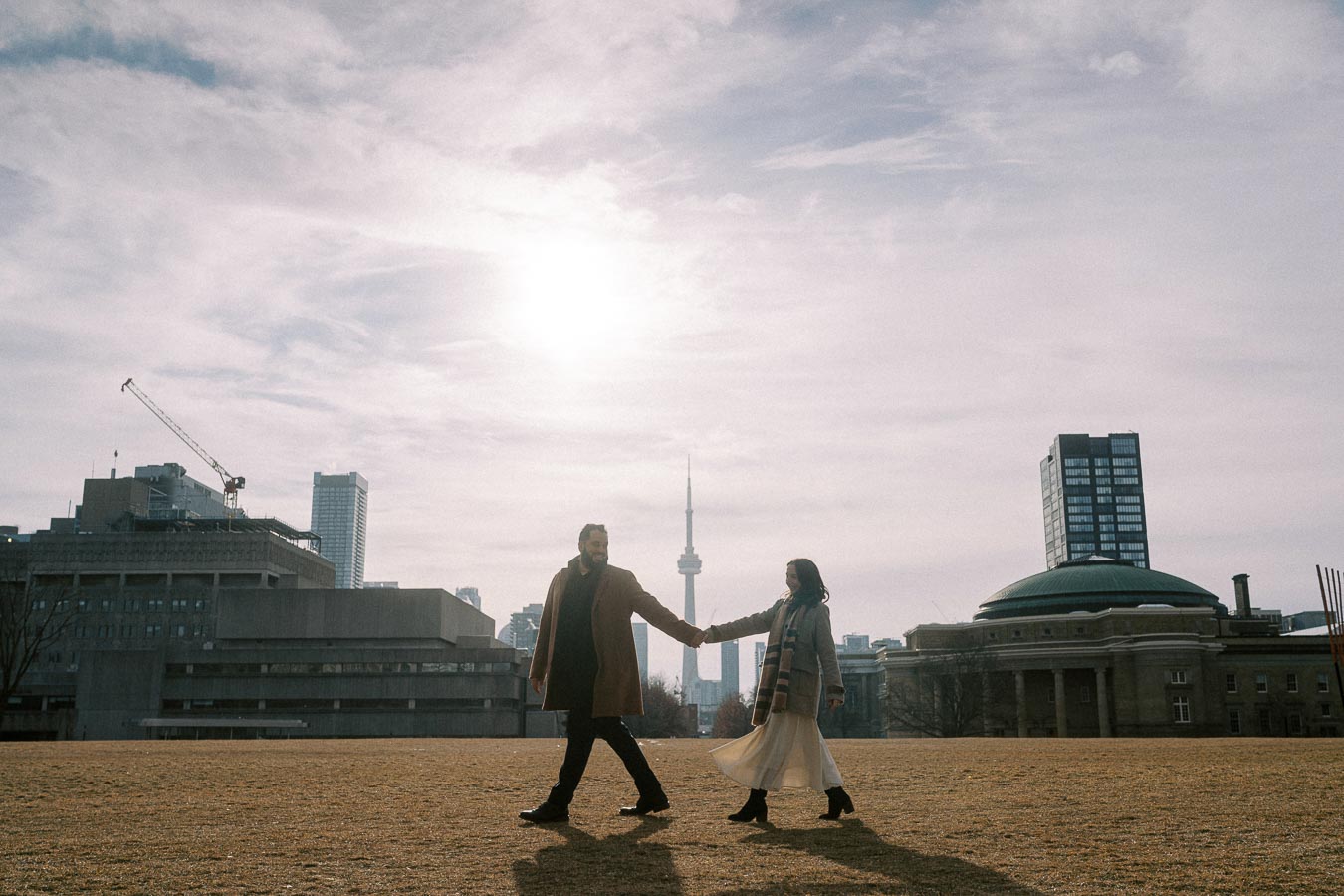 Couple holding hands walking in an open field with Toronto skyline and CN Tower in the background under a cloudy sky.