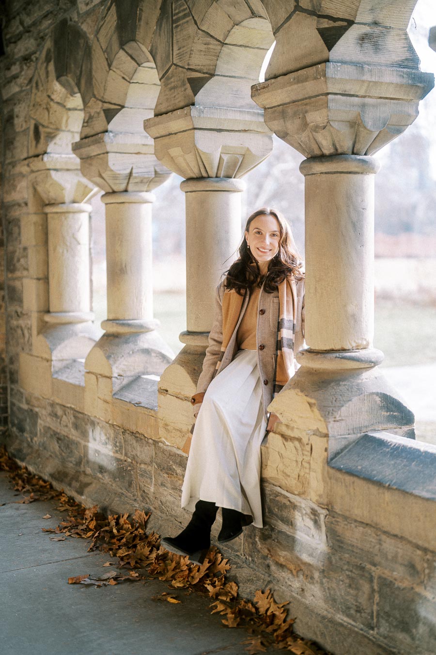 Young woman smiling while sitting on a stone ledge in a historic, arched corridor, wearing a beige coat and white dress with fallen leaves around.