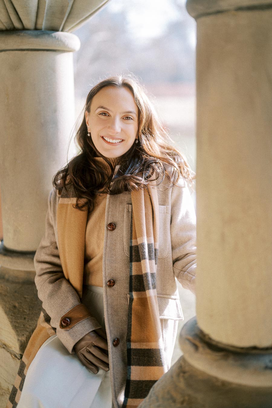 A woman smiling in a stylish autumn outfit, wearing a cozy coat and plaid scarf, standing between stone columns with soft natural lighting.