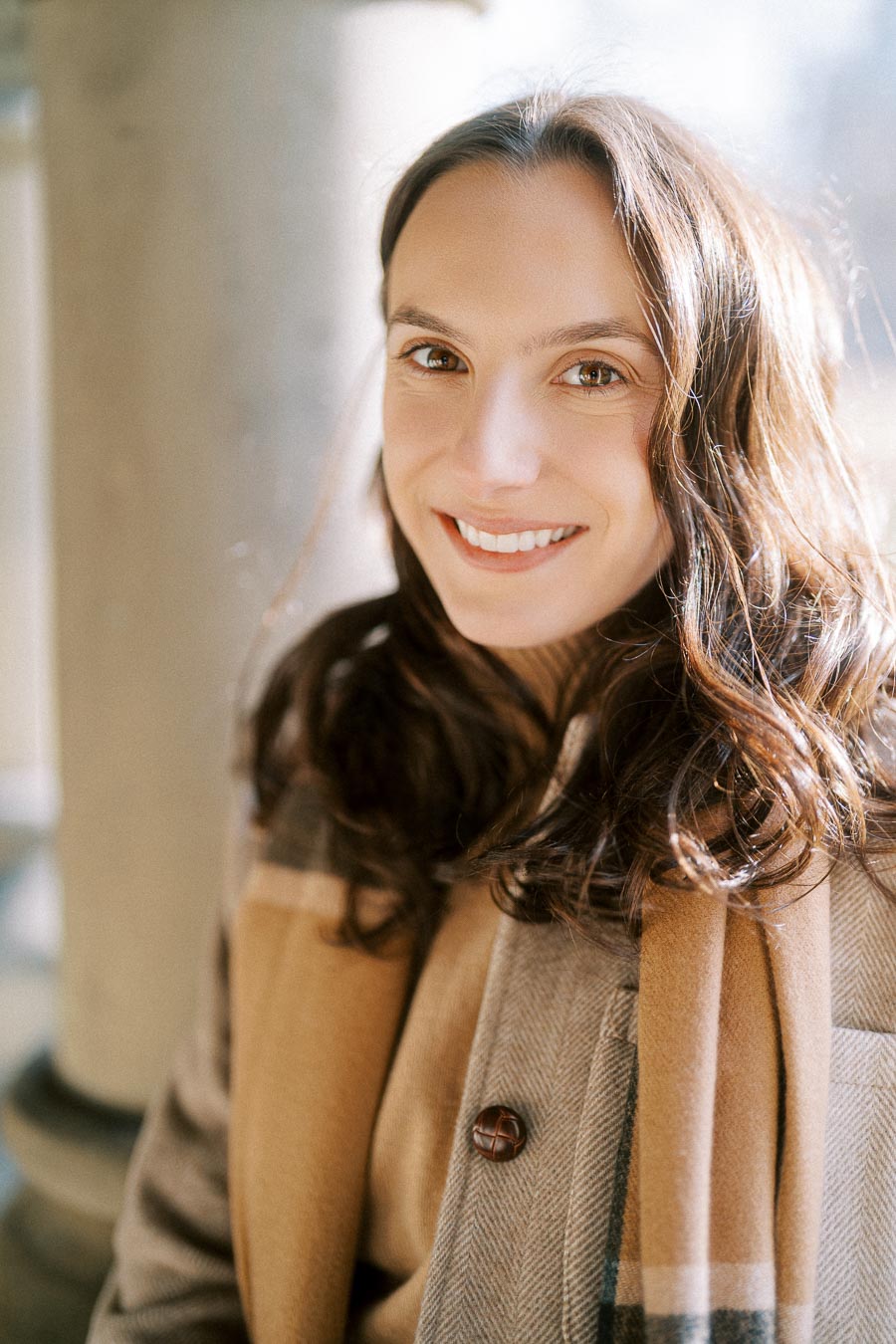 Smiling woman with long brown hair wearing a plaid coat and scarf in natural light.
