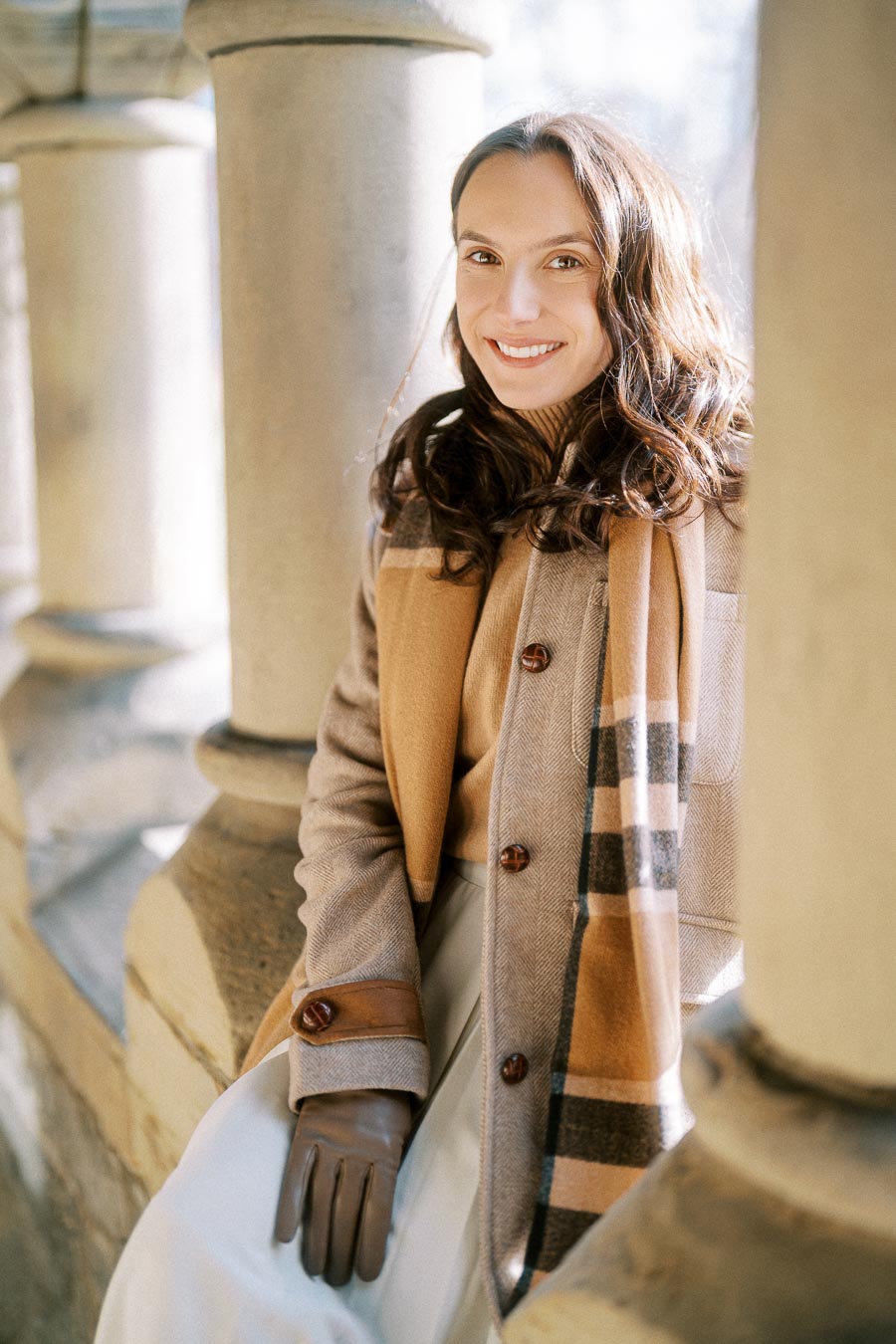 Smiling woman in a stylish winter coat and scarf sits by stone columns, sunlight highlighting her brown hair and elegant outfit.