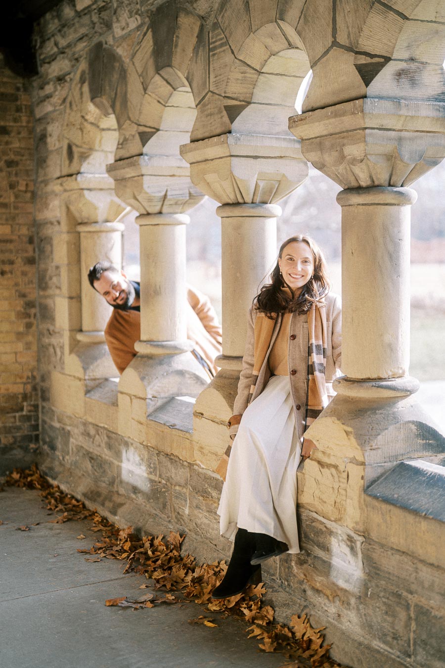 A woman sitting on a stone ledge with autumn leaves on the ground, wearing a beige coat and scarf, smiling in a sunlit, arched stone corridor. A man playfully peeks through the arches in the background.
