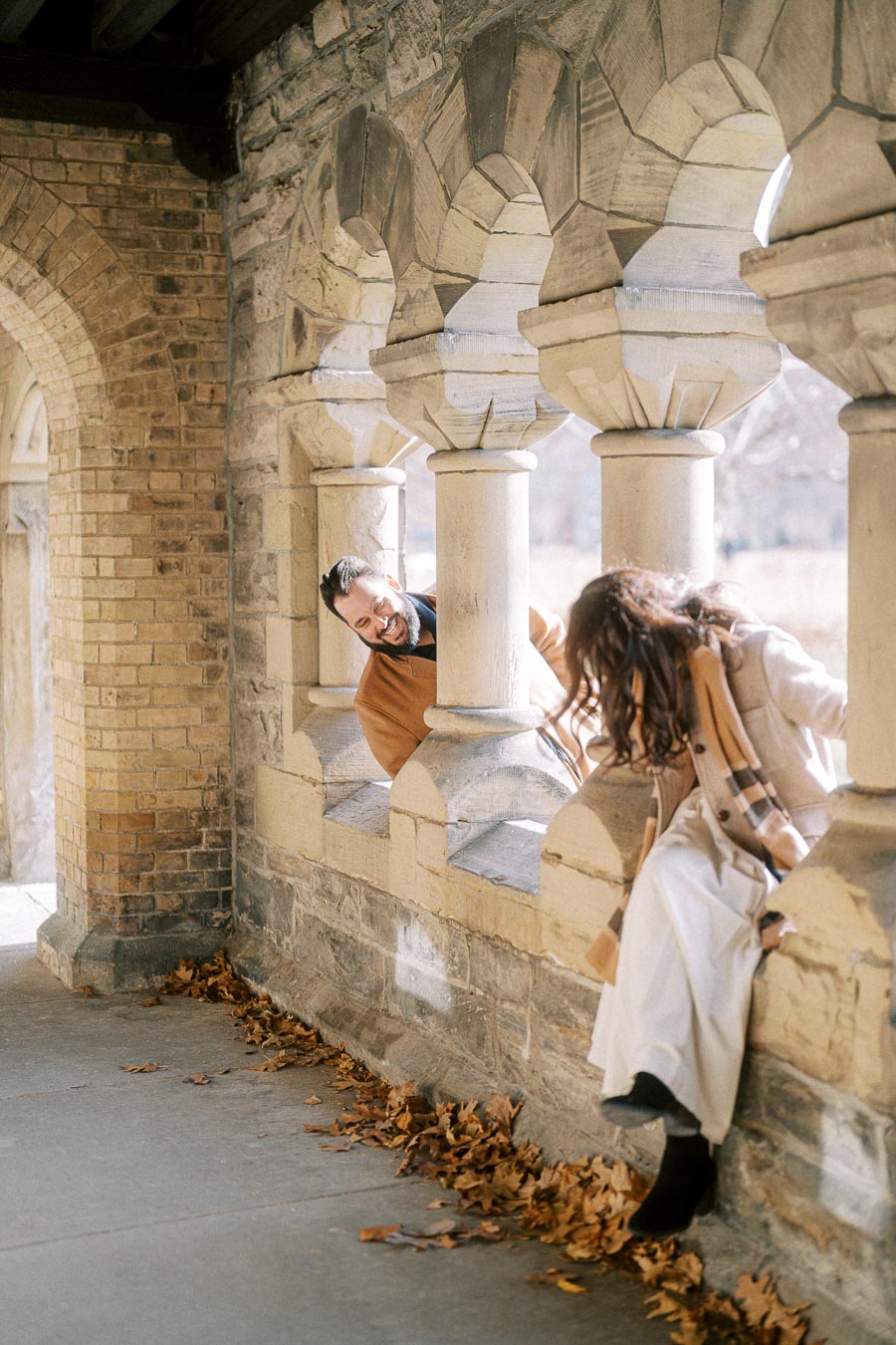 A woman sitting playfully on a stone ledge flanked by arched pillars, with a man peeking through the column next to her, both smiling and enjoying a sunny autumn day surrounded by fallen leaves.