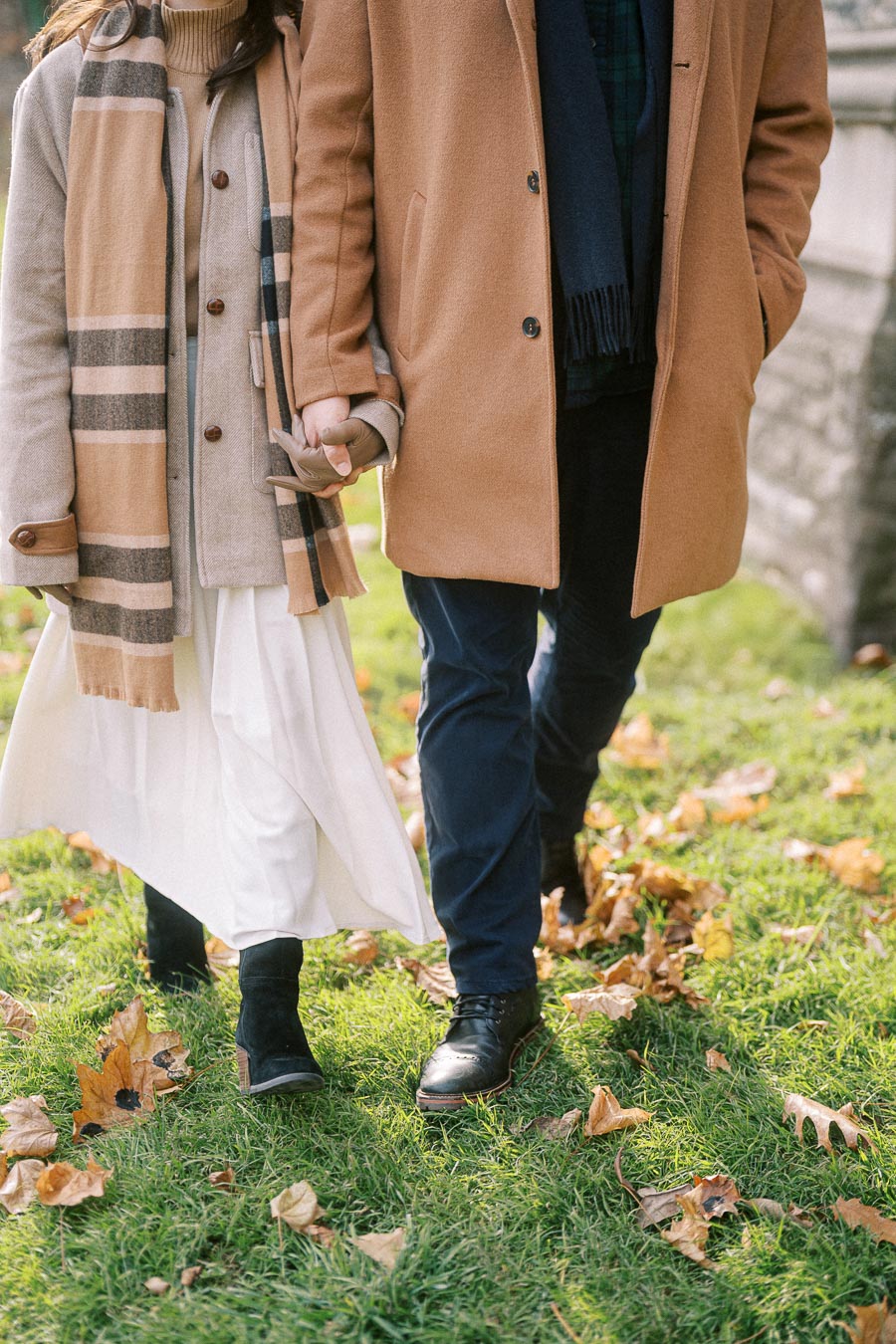 A couple holding hands and walking on a grassy park path covered with autumn leaves, dressed in stylish beige and tan coats, symbolizing romance and fall fashion.