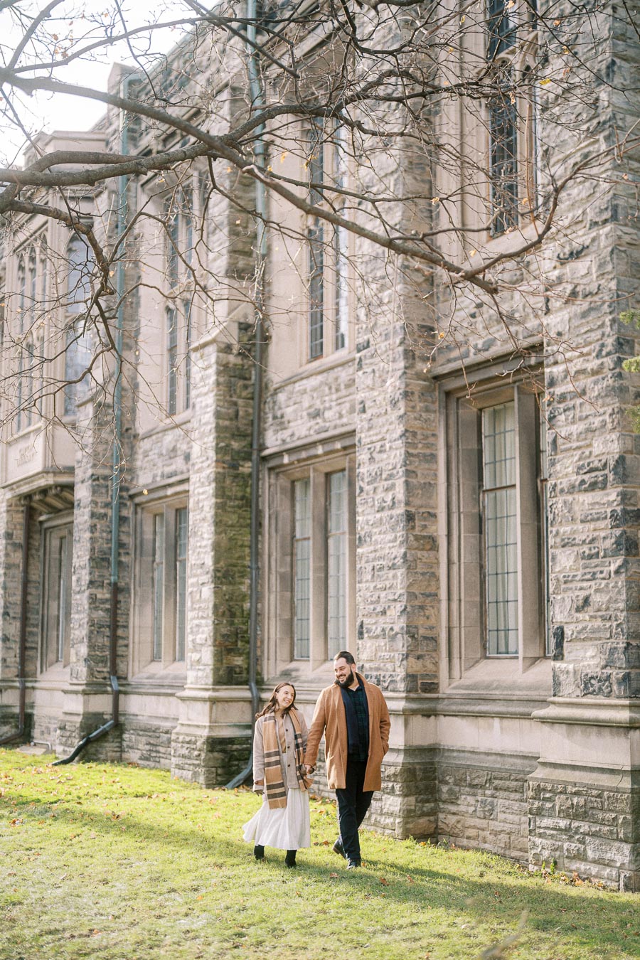 A couple walking hand in hand beside a historic stone building on a sunny day, wearing winter coats and scarves.