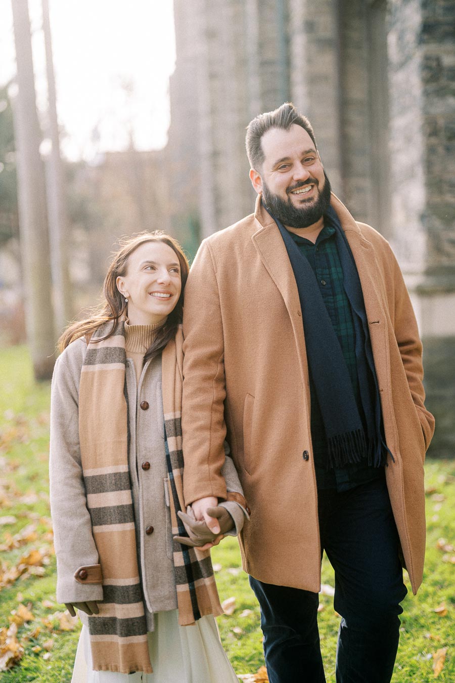 A happy couple walking outdoors, wearing matching beige coats and scarves, enjoying a fall day.