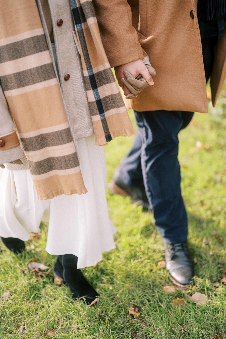 A couple in stylish winter clothing walking hand in hand on a grassy path, wearing camel-colored coats and scarves.