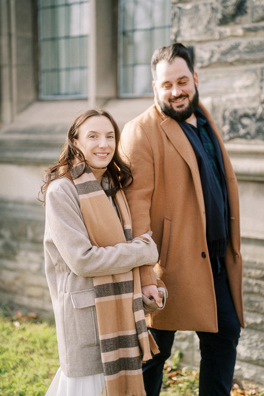 Couple wearing fall coats and scarves walking hand-in-hand outside a historic stone building, showcasing cozy autumn fashion.
