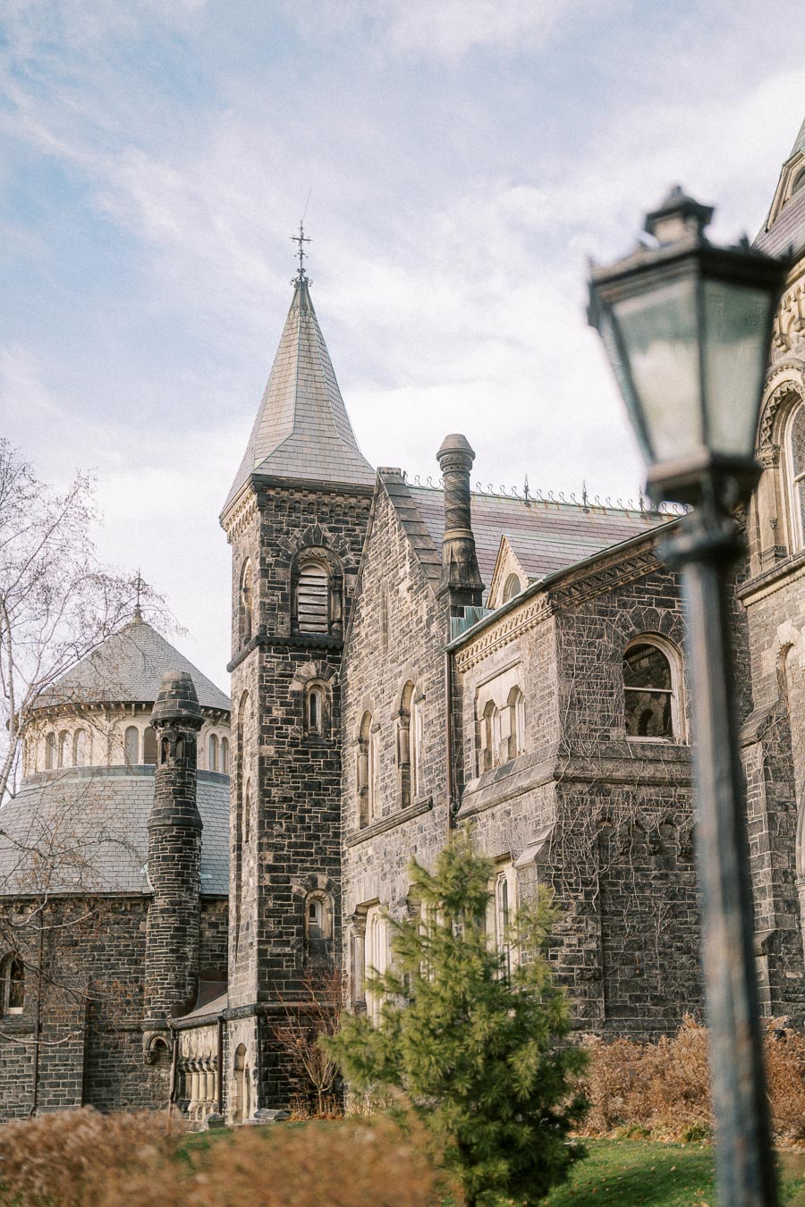 Historic stone Gothic building with tall spires and arched windows under a clear blue sky, framed by a vintage street lamp in the foreground and lush greenery in the background.