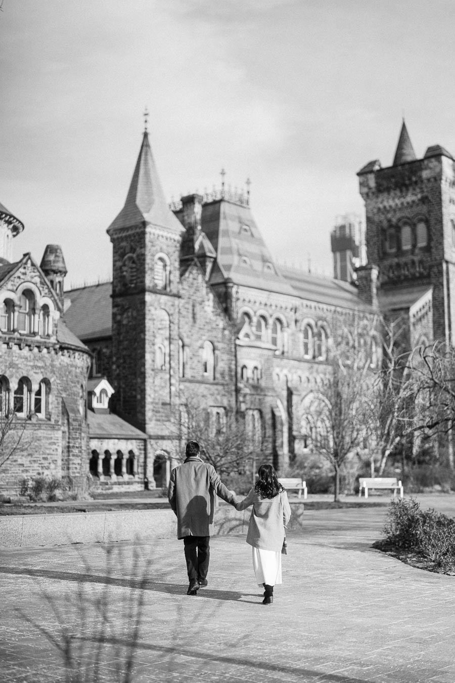 Black and white photo of a couple walking hand in hand towards a historic, Gothic-style building on a clear day, with leafless trees and empty benches lining the pathway.