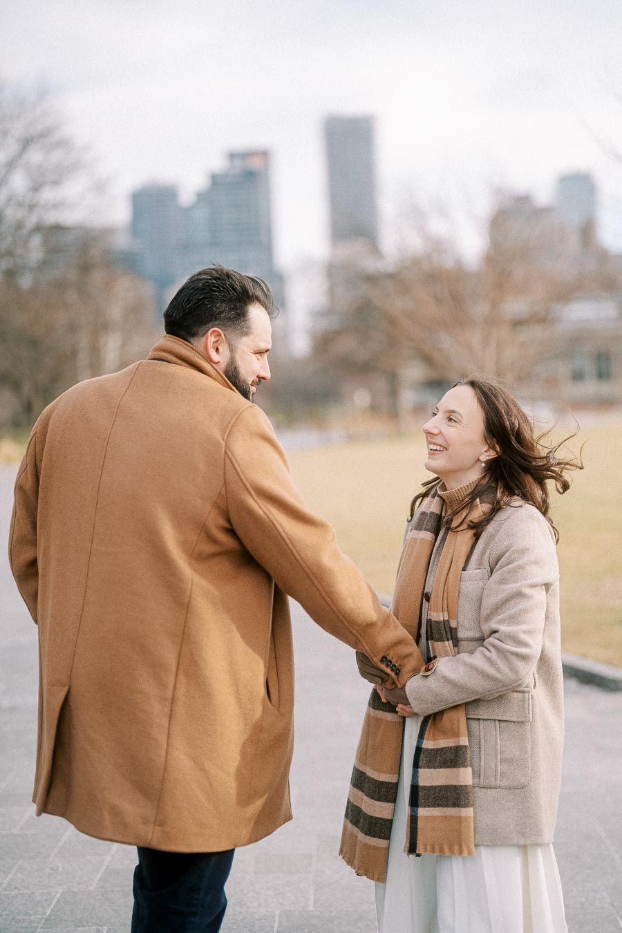 Couple smiling and holding hands in a city park on a winter day, wearing warm coats and scarves, with urban skyline in the background.