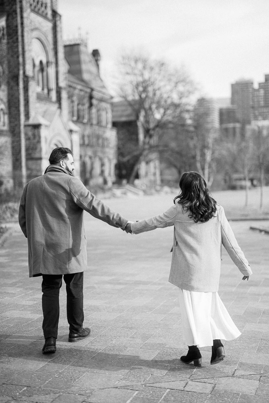 A black and white photo of a couple walking hand in hand down a cobblestone path, with historic buildings in the background, showcasing a romantic scene in an urban setting.