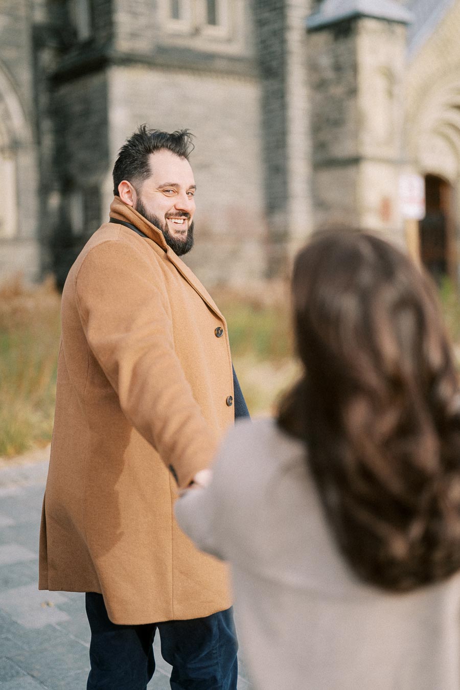A man in a tan coat smiles while holding hands with a woman in front of a historic stone building, creating a warm and romantic atmosphere.