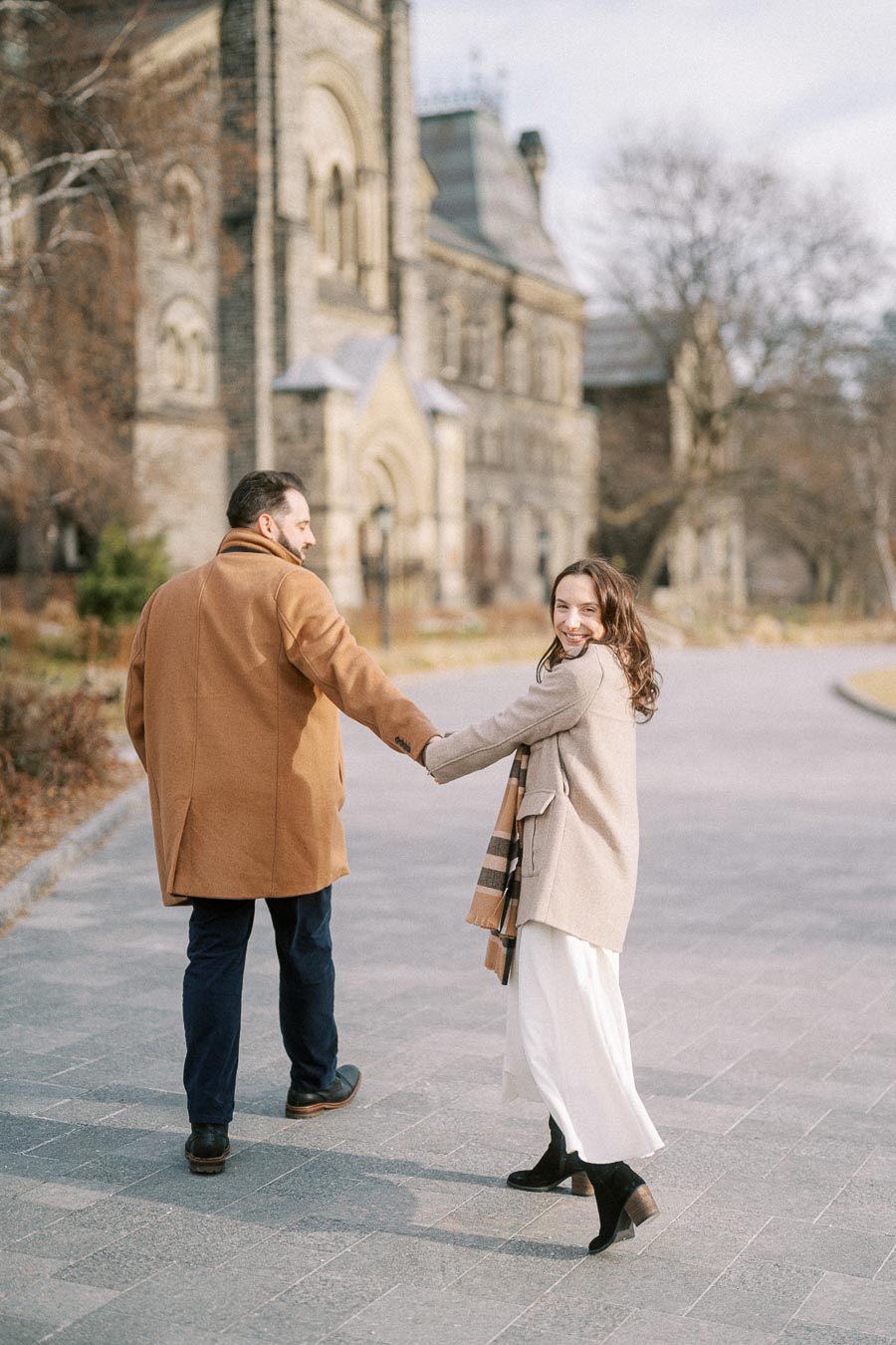 A couple walking hand in hand near a historic building, with the woman smiling back at the camera wearing neutral-toned coats on a sunny day.