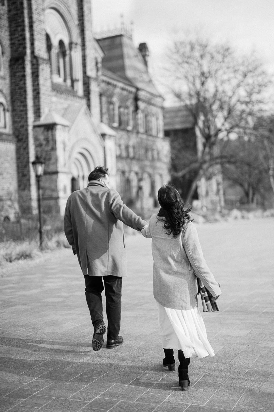 Black and white image of a couple walking hand in hand along a historic cobblestone path, bordered by an old stone building with arched windows, conveying romance and nostalgia in an urban setting.