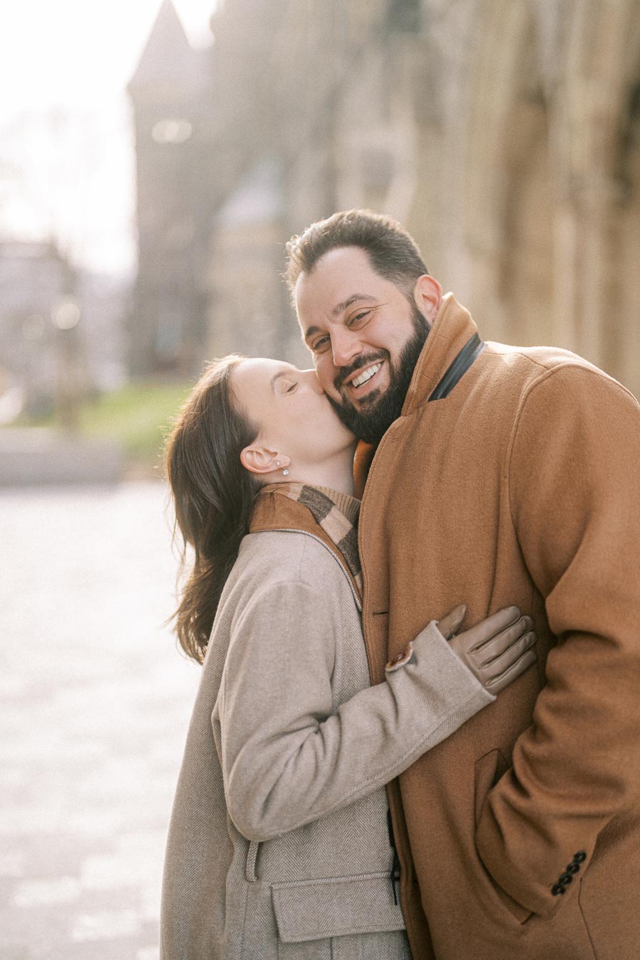 Happy couple embracing outdoors in cozy winter coats, with woman lovingly kissing man's cheek, city background softly blurred.