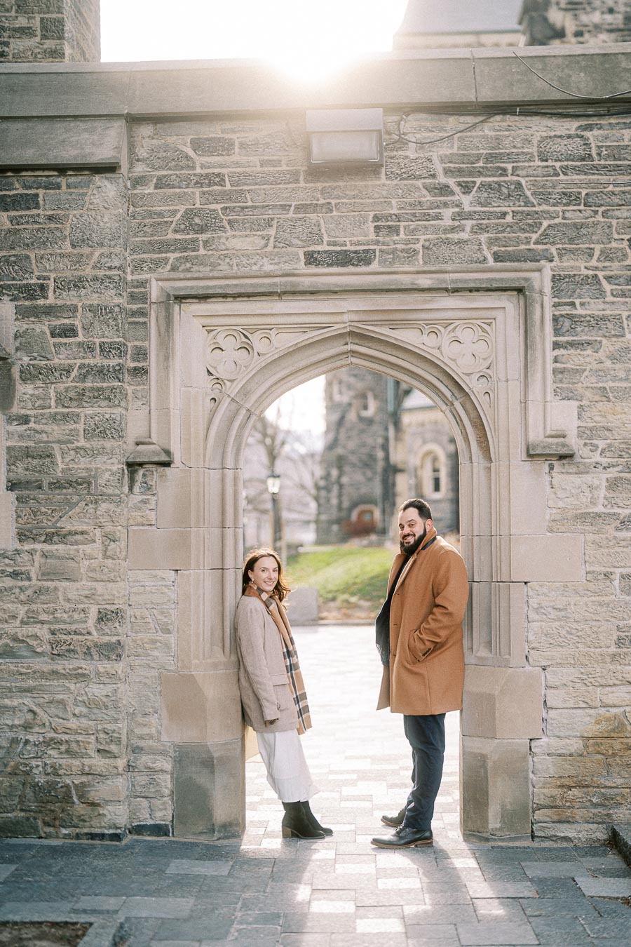 A stylish couple poses under a stone archway in a scenic courtyard, wearing elegant coats and scarves, with sunlight peeking over the historic building.