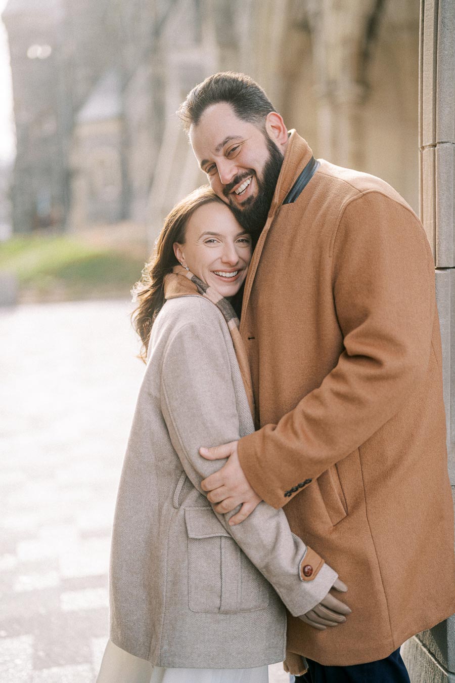 A happy couple embracing in stylish coats, standing outside on a sunny day with a historic building in the background.