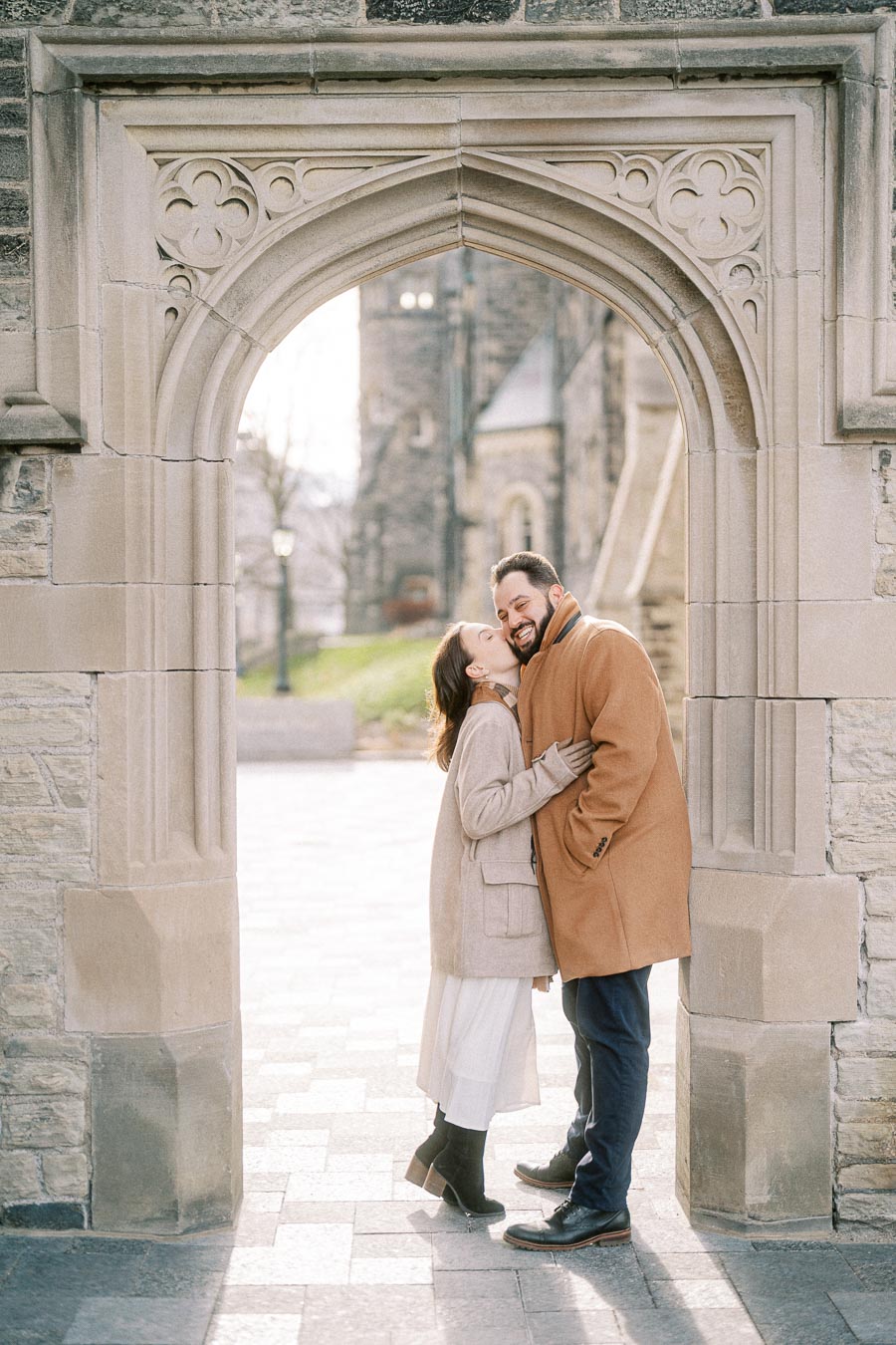 A couple in winter coats shares a joyful moment under a stone archway in a historic setting.
