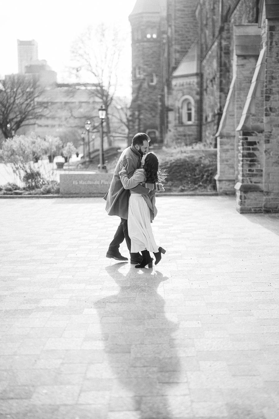 A black and white photo of a couple dancing joyfully on a sunlit cobblestone path, with historic architecture in the background.