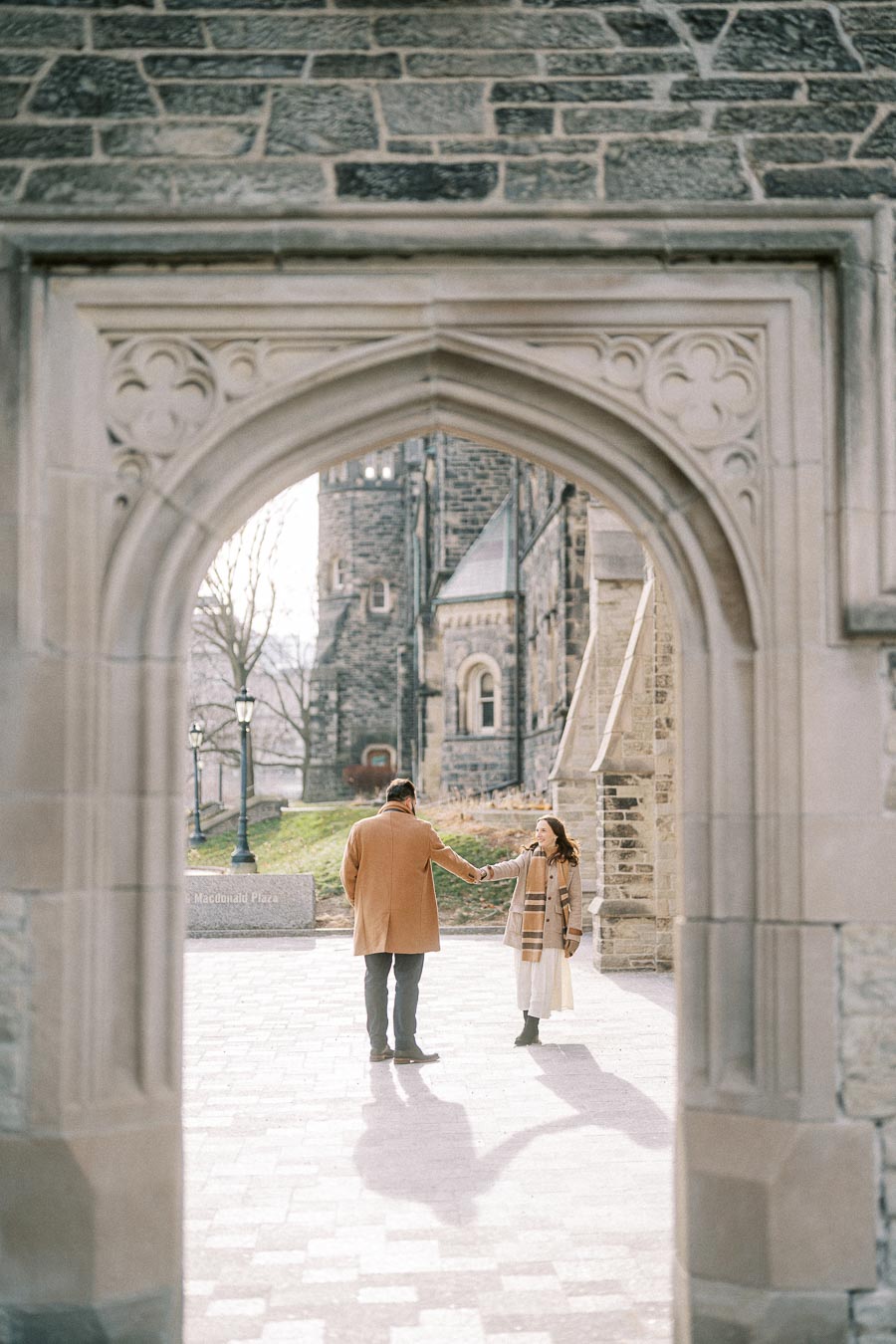 A couple holding hands while standing in a historic stone archway, showcasing a romantic scene at a university campus.