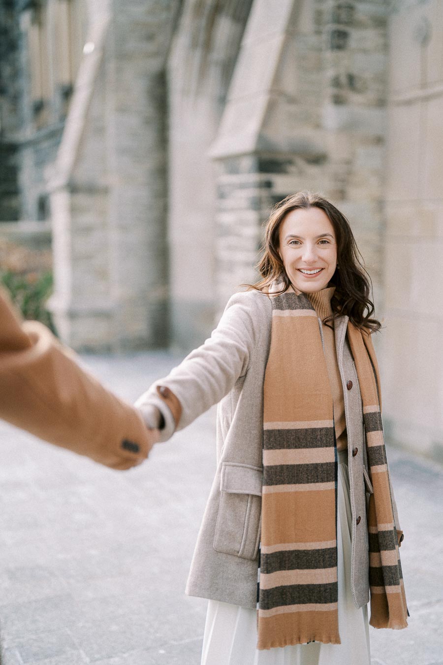 Woman in a beige coat and scarf smiling while holding someone's hand, standing in front of a stone building.