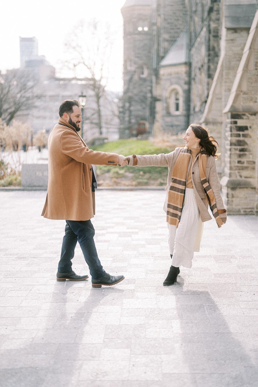 A couple enjoying a sunny day outdoors in stylish winter coats, smiling and holding hands in front of a historic stone building.