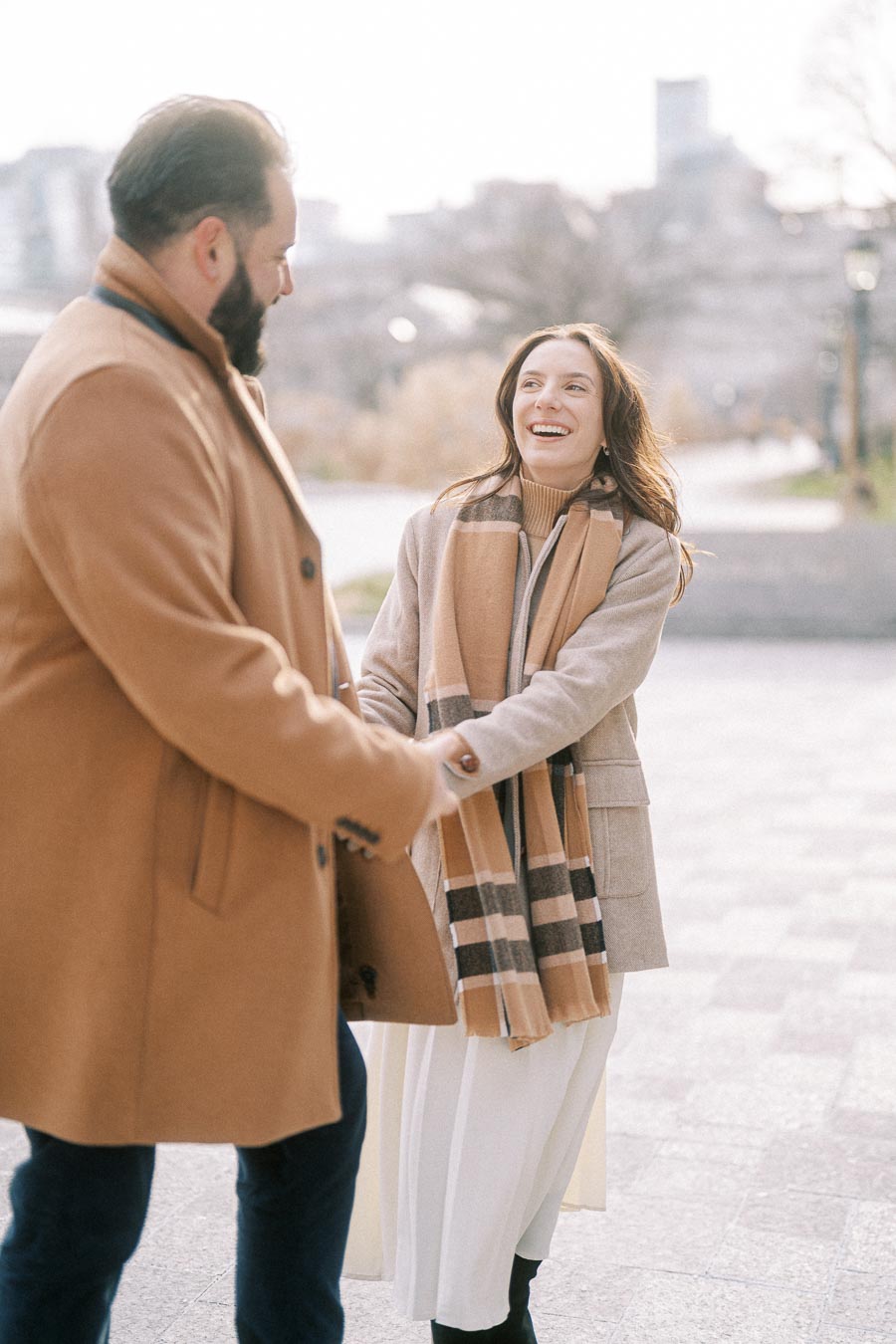 A happy couple walking in the park on a sunny day, wearing stylish winter coats and scarves, with city buildings in the background.