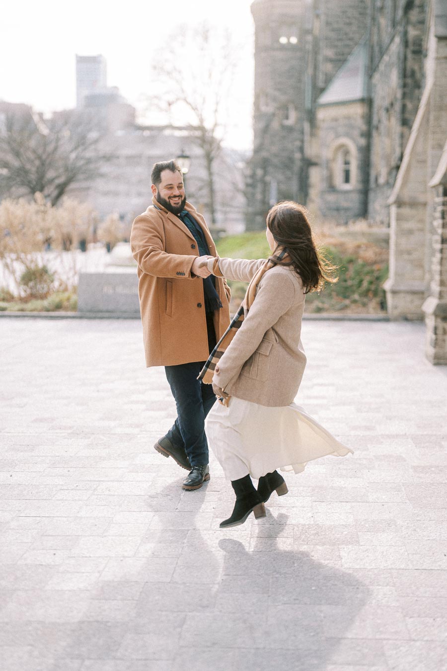 Happy couple dancing outside a historic stone building, both wearing stylish winter coats and smiling joyfully.