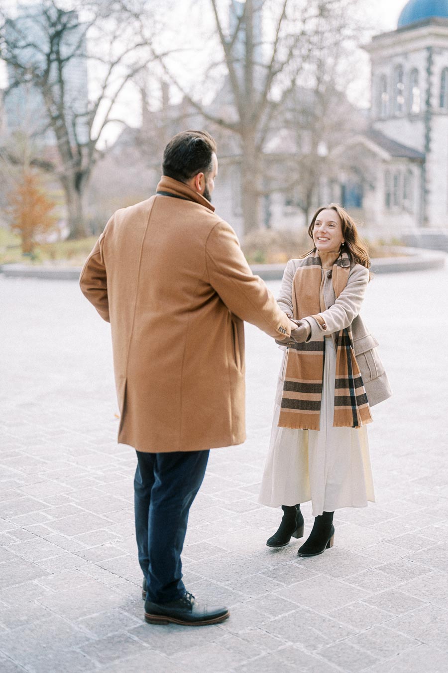 A couple holding hands and smiling in a scenic, winter urban setting with trees and historic architecture in the background, both wearing stylish winter coats and scarves.