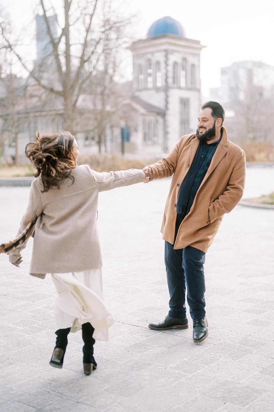 A couple joyfully dancing outdoors in a park setting, both wearing stylish winter coats. The man smiles warmly as they twirl, with a historic building in the background creating a picturesque winter scene.