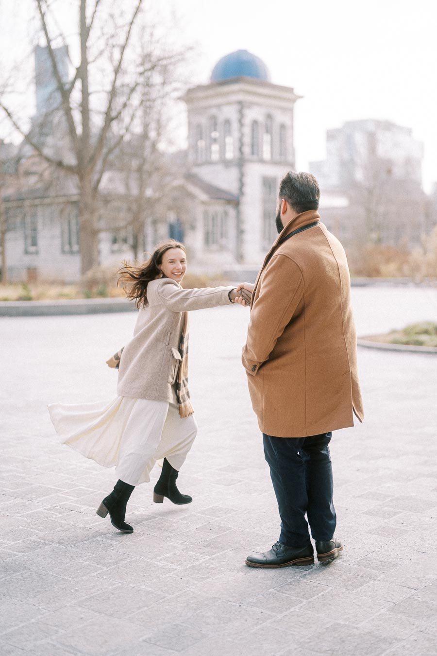 A woman in a beige coat joyfully twirls while holding hands with a man in a brown coat, in front of a historic building with a blue dome, creating a lively urban scene.