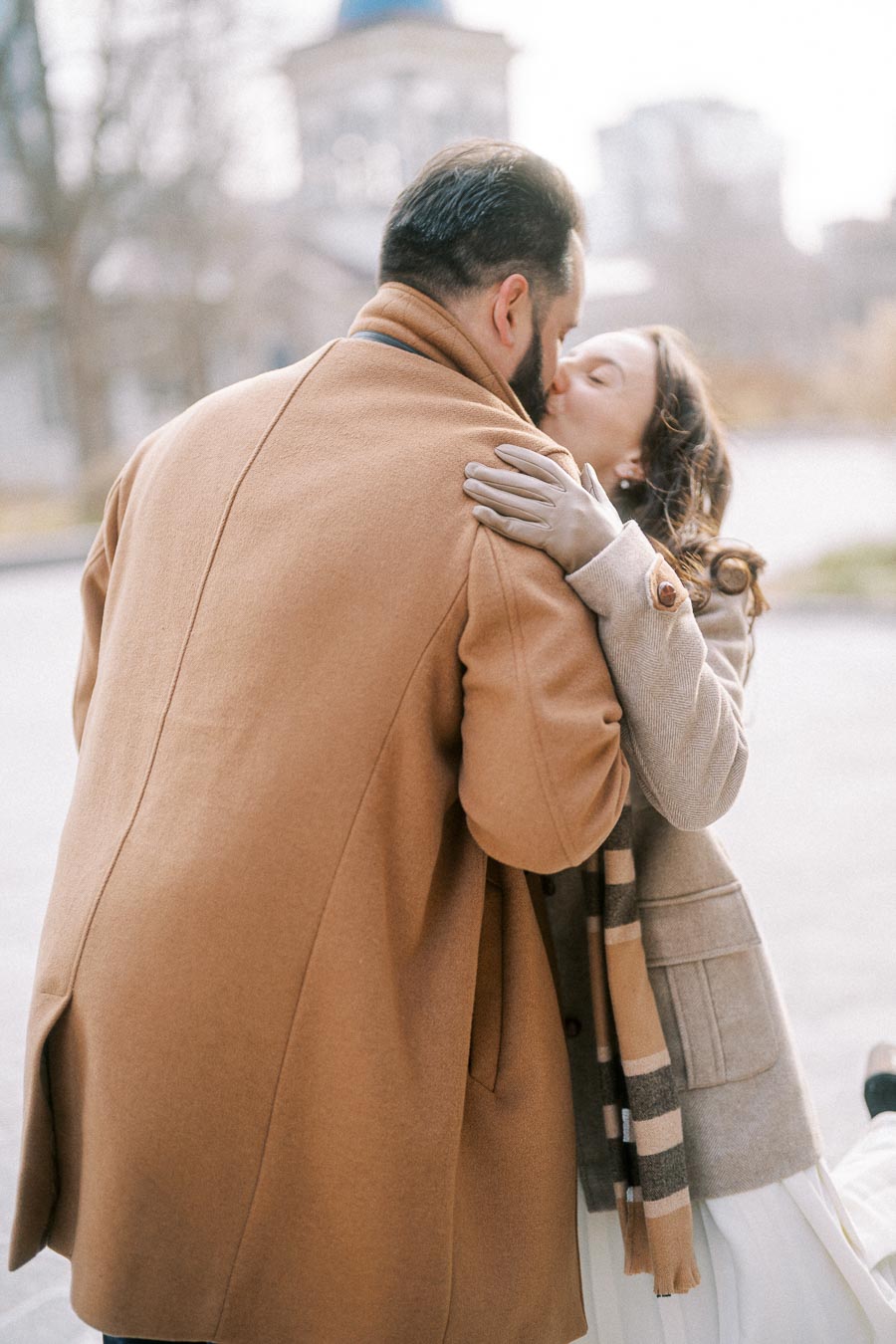 A couple sharing a tender kiss on a chilly day, both dressed in warm winter coats, with a blurred architectural background.