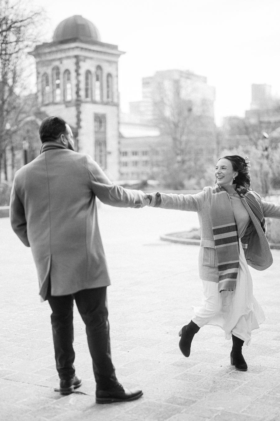 A couple joyfully holding hands and spinning in a scenic urban setting, with historic architecture in the background. The woman is wearing a scarf and a long coat, while the man wears a similar coat, creating a romantic atmosphere. Black and white photo, capturing a moment of happiness.