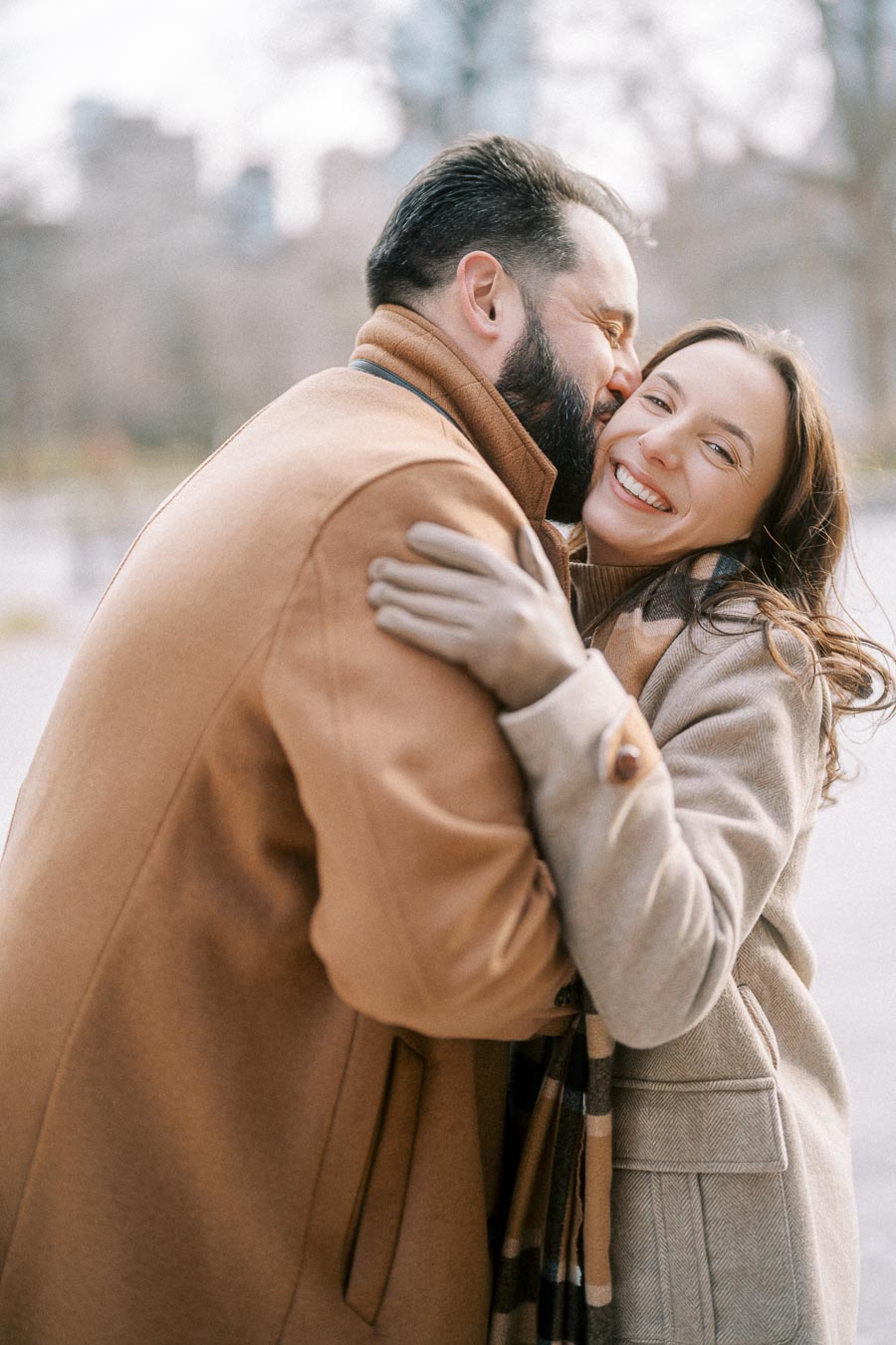 A happy couple embraces outdoors, wearing warm coats in a winter setting with blurred city buildings in the background, conveying warmth and affection.