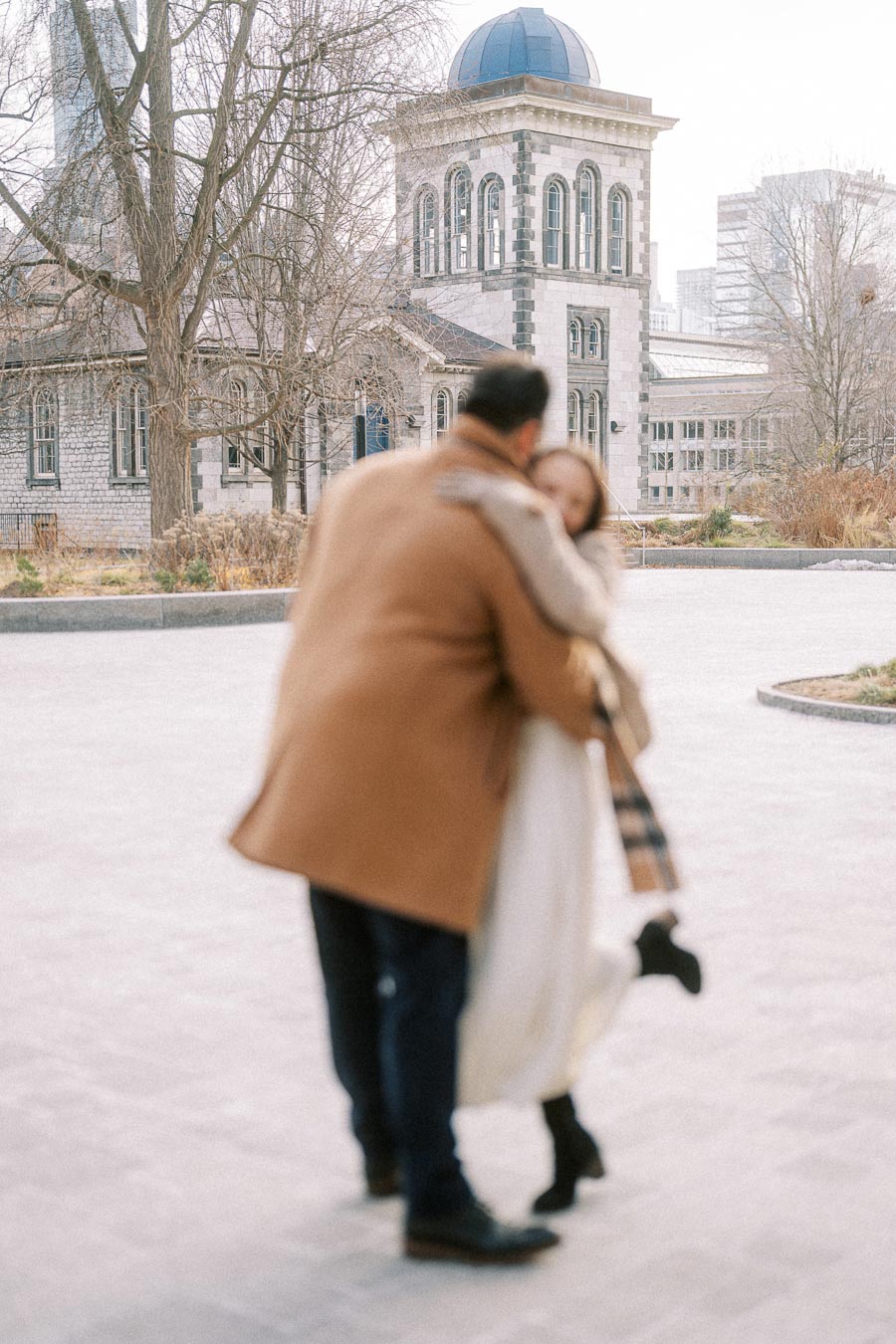 A blurred image of a couple embracing in a park setting with a historical stone building and bare trees in the background. The man is wearing a brown coat and the woman is in a white outfit with a plaid scarf. Urban architecture and winter ambiance.