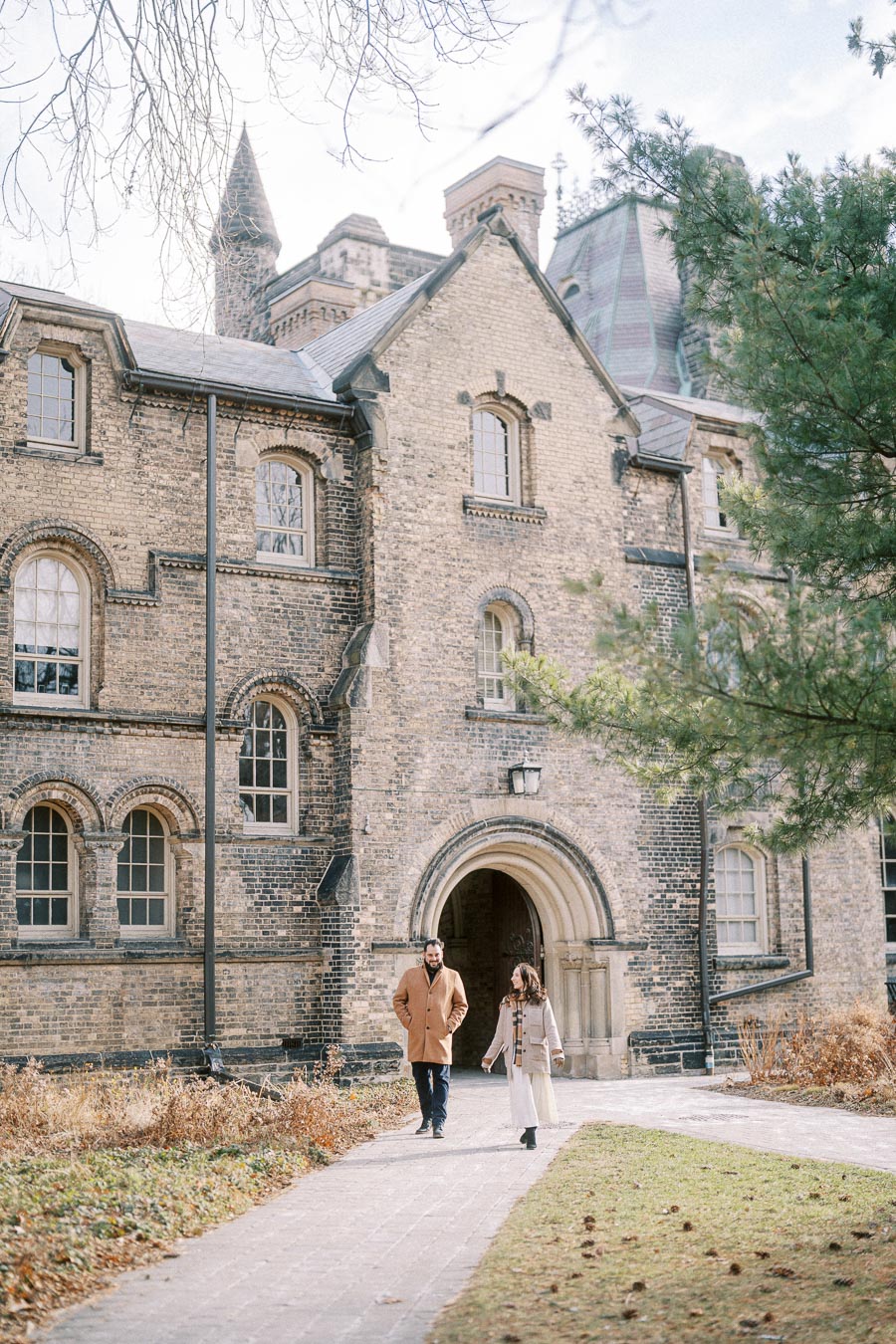 Couple walking along a pathway in front of a historic brick building with arched windows and a pointed tower, surrounded by greenery and clear skies.