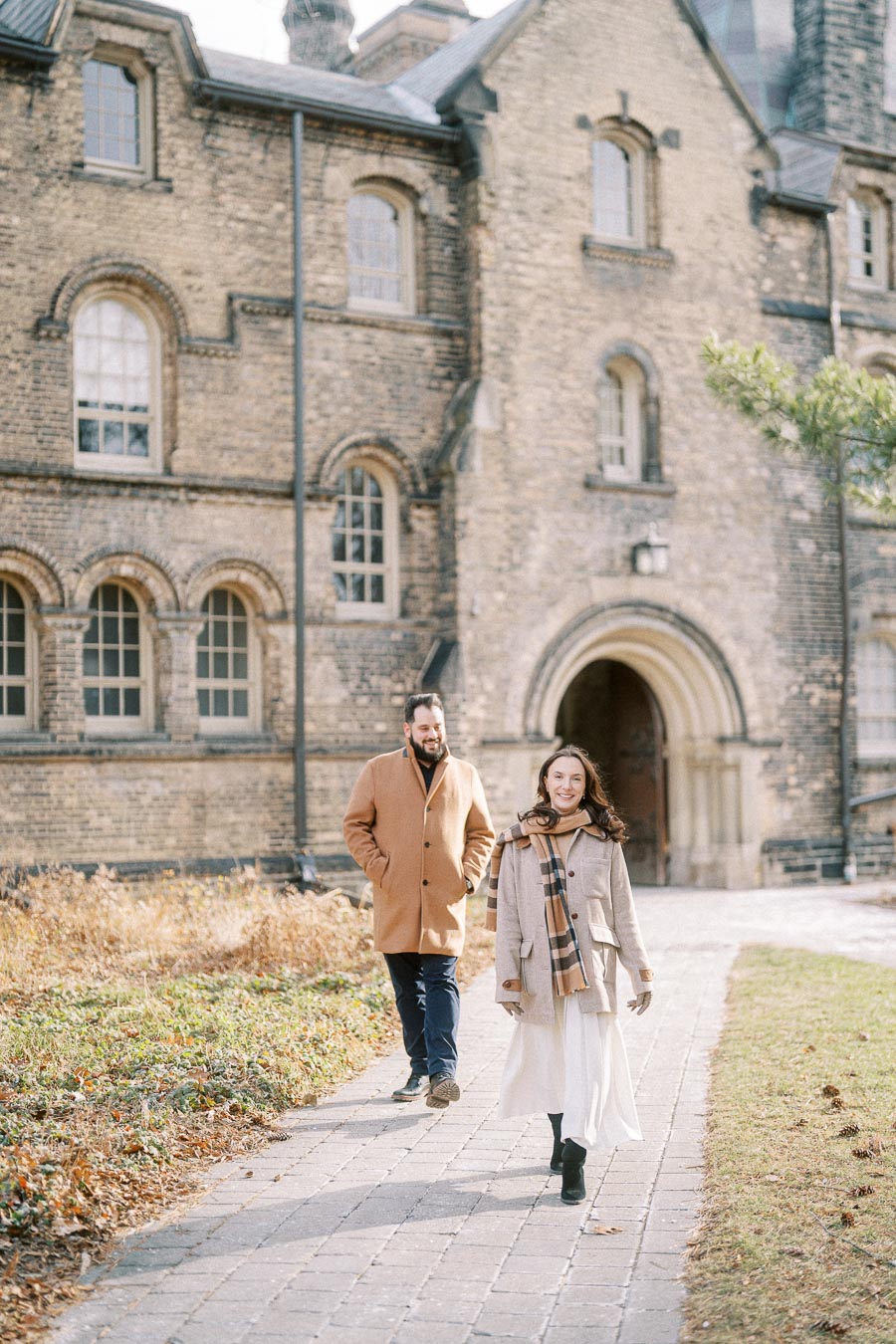A couple dressed in stylish winter coats walking along a cobblestone path in front of an old brick building with arched windows, capturing a romantic fall or winter scene.