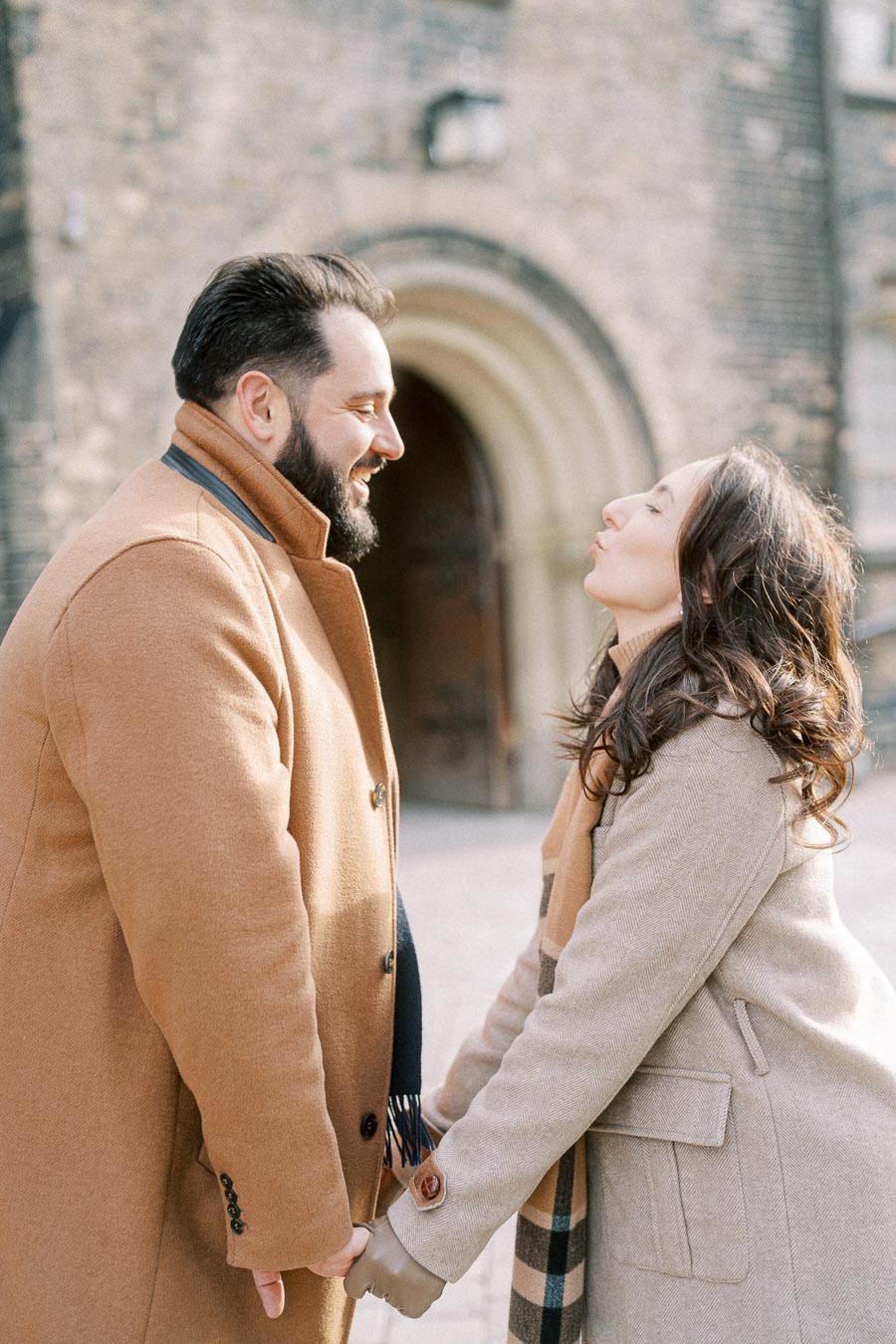 A couple in warm coats smiling and holding hands in front of a historic stone building entrance.