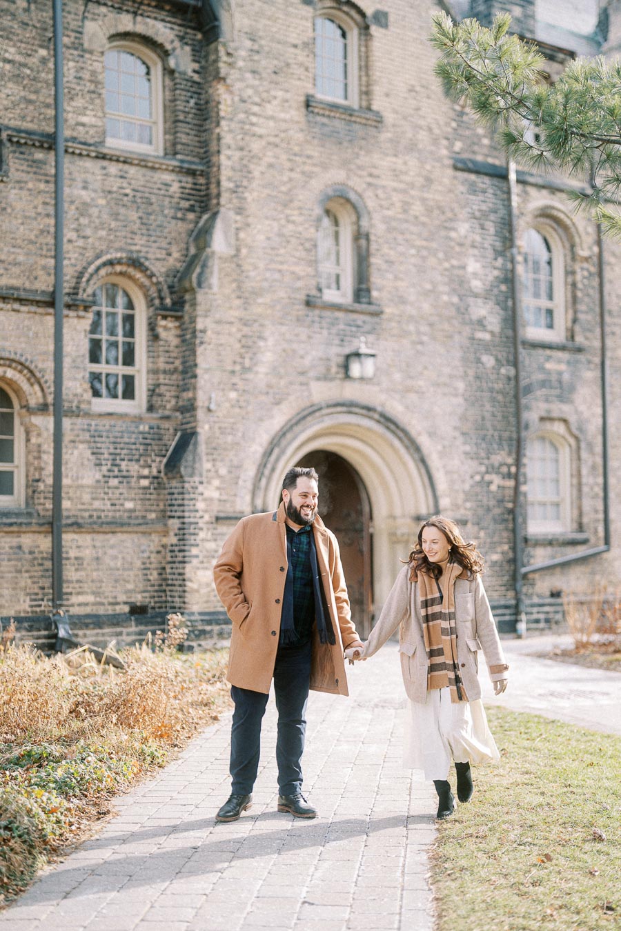 A couple walking hand in hand on a sunny day in front of a historic brick building with arched windows.