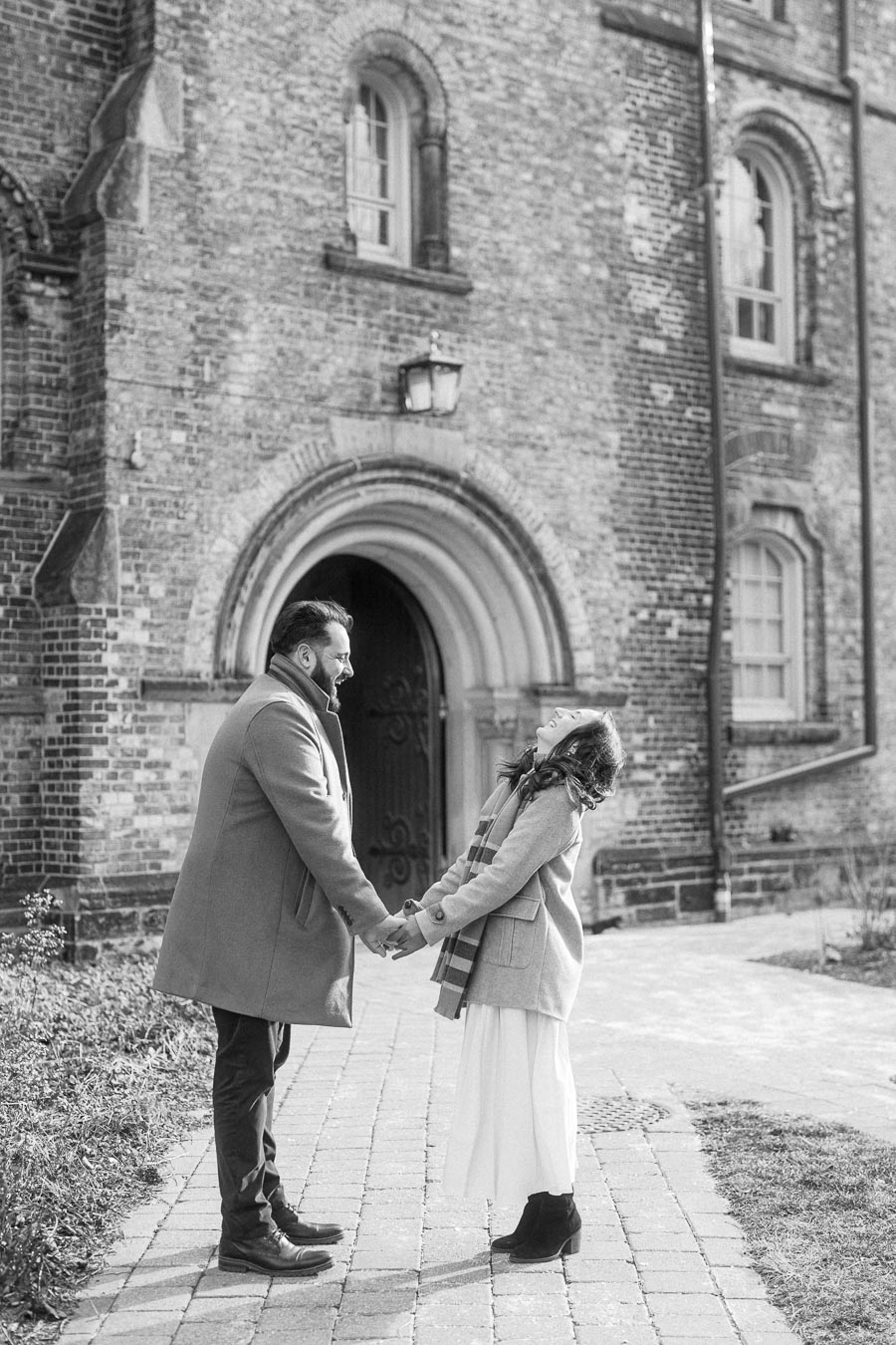A couple joyfully holding hands outside a historic brick building, expressing happiness and connection in a black and white photograph.