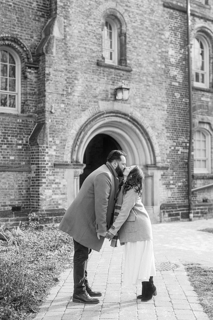A couple shares a loving kiss in front of a historic brick building, embracing on a cobblestone path.