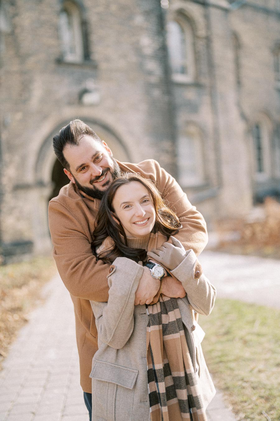 A smiling couple in warm coats embracing outdoors on a sunny day, with a historic stone building in the background.