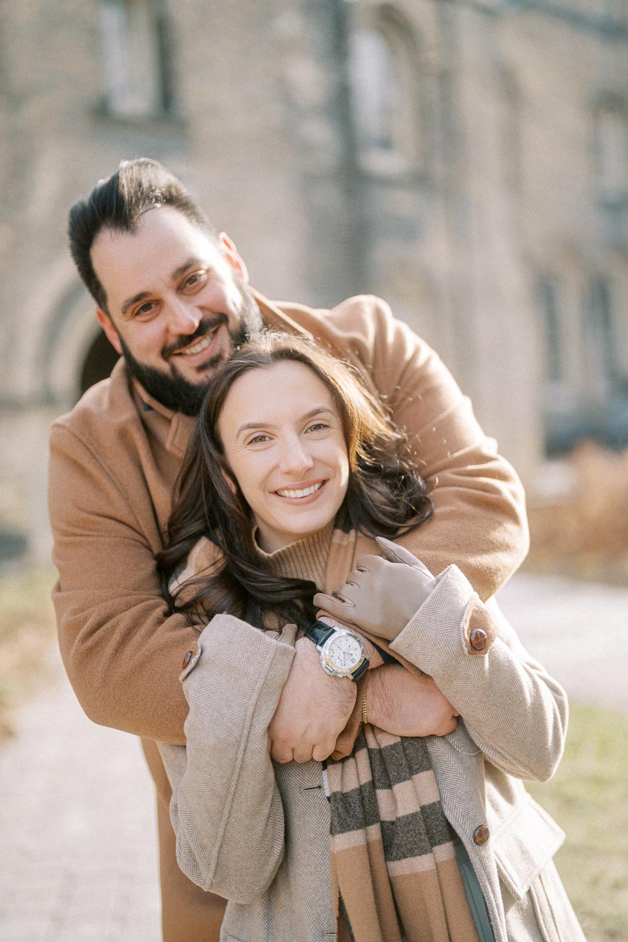 A happy couple embracing outdoors on a sunny day, both wearing stylish winter coats, smiling warmly at the camera, with a stone building in the background.