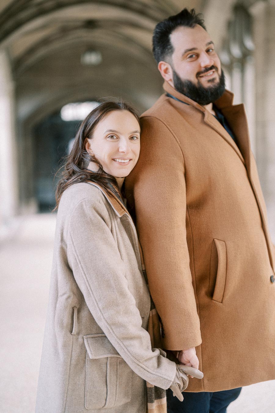 A smiling couple holding hands, dressed in stylish winter coats, standing in a beautifully lit corridor showcasing architectural details.
