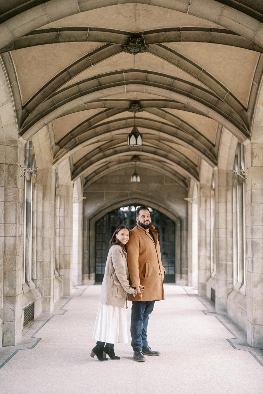 A couple in stylish winter coats standing in an ornate stone archway, holding hands and smiling.